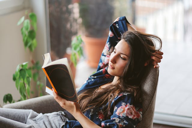 A woman in a floral shirt relaxes in a chair, reading a book. She has long hair and appears absorbed...