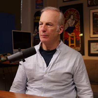 Bob Odenkirk sits at a table in an interview setting, wearing a light-colored shirt. A microphone and drink are in front of them, with colorful posters on the walls.