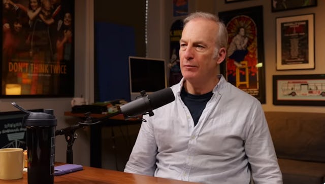 Bob Odenkirk sits at a table in an interview setting, wearing a light-colored shirt. A microphone an...