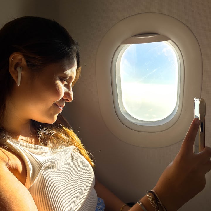 A young woman sitting by an airplane window holds a phone in one hand, smiling softly as sunlight il...