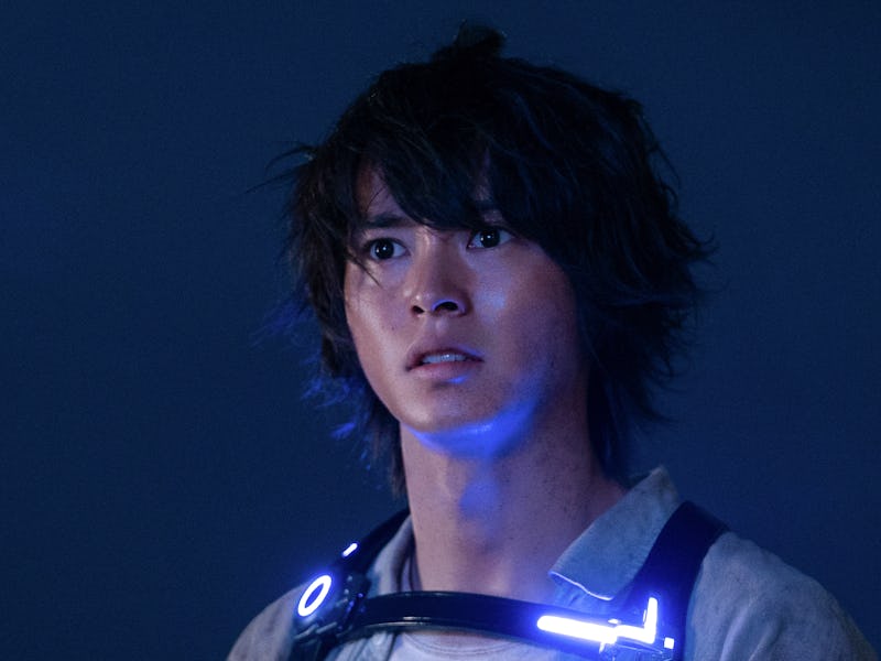 A young man with tousled hair and an expression of concern stands against a dark background, wearing...
