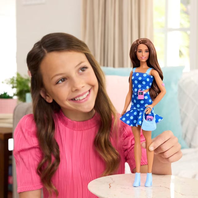 A smiling girl in a pink shirt holds a doll wearing a blue polka dot dress. They are seated in a coz...