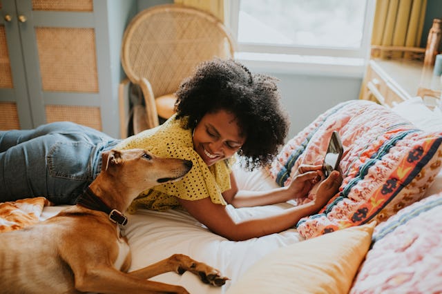 A smiling woman with curly hair lies on a bed, playfully interacting with a dog while holding a tabl...