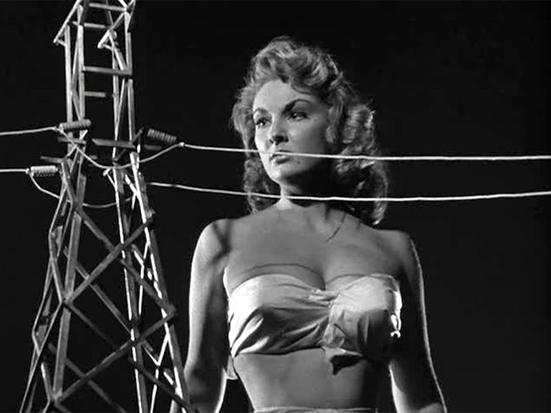 A black-and-white image of a woman in a strapless top standing near a tall power line tower, with po...