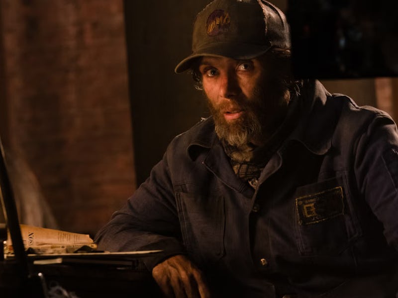 A rugged man with a beard and mustache, wearing a cap and work jacket, sits at a table. Books and pa...