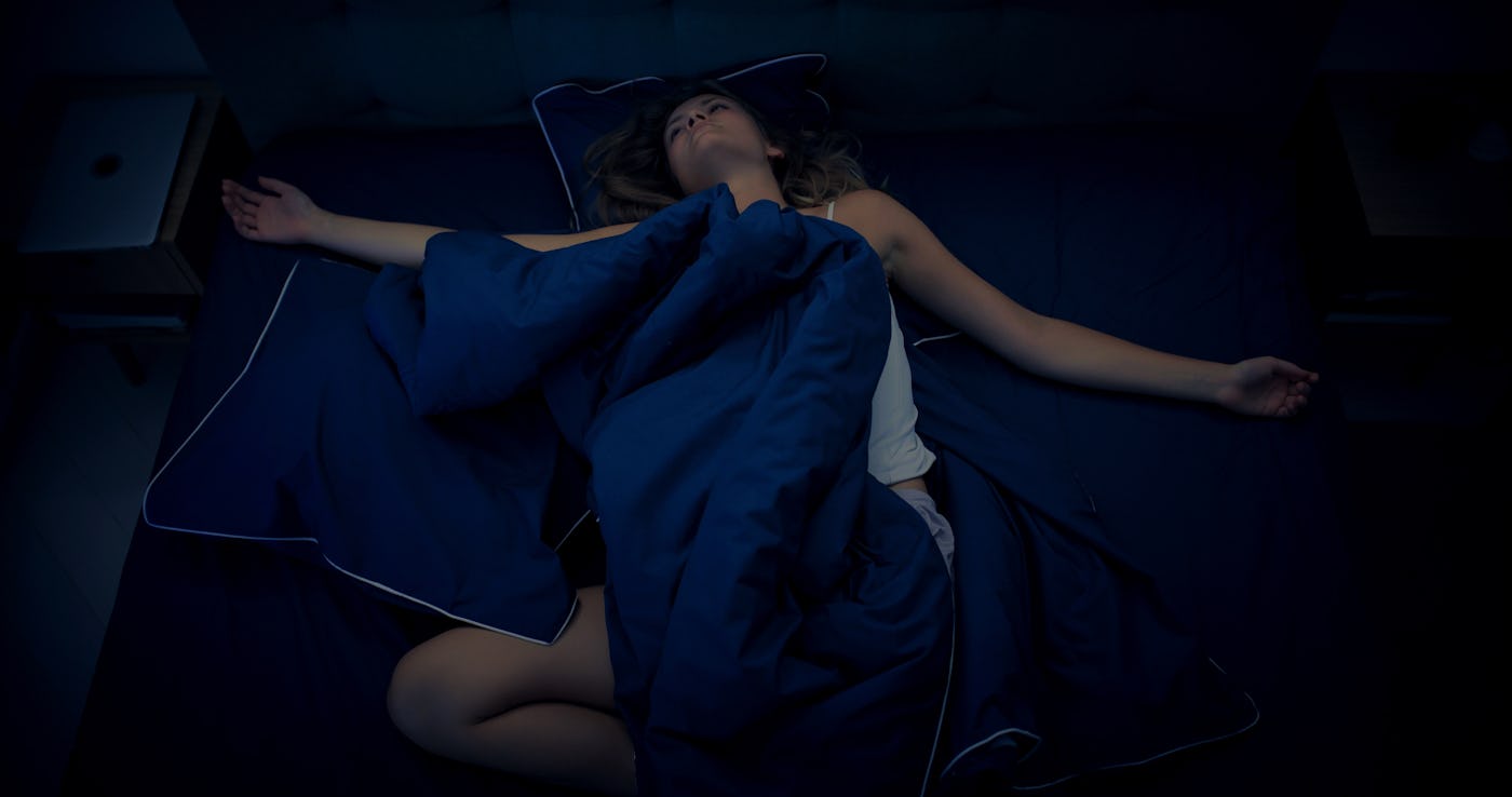 A woman lies on a dark bed, stretched out under a blue blanket. Her arms are open, and she appears relaxed, surrounded by a calm, dim atmosphere.