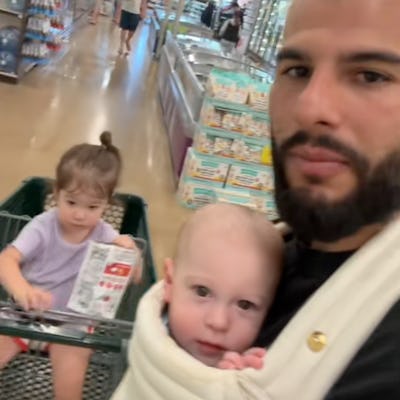 A man with a beard holds a baby in a carrier while a toddler sits in a shopping cart. They are in a grocery store aisle with shelves lined with products.