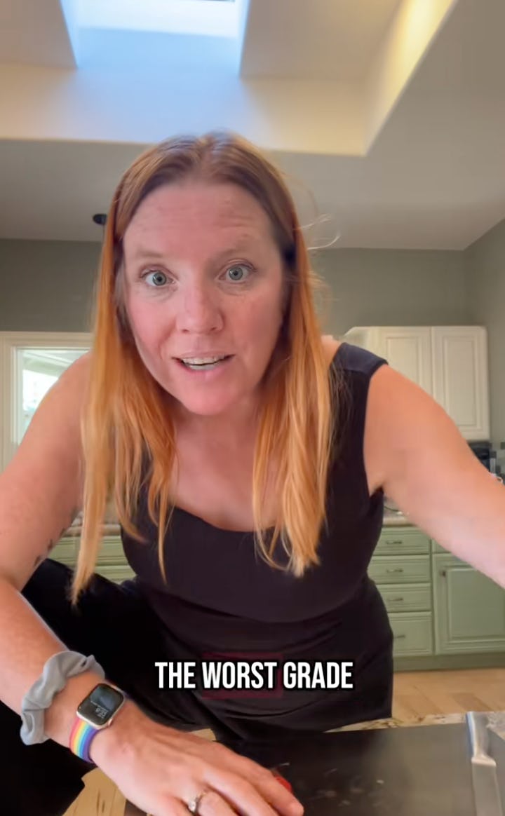 A woman with long red hair smiles while leaning over a table, looking surprised. Text below her reads, "THE WORST GRADE." A kitchen decorates the background.
