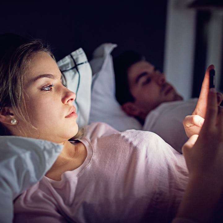 A woman scrolls on her phone in bed as her husband sleeps beside her.