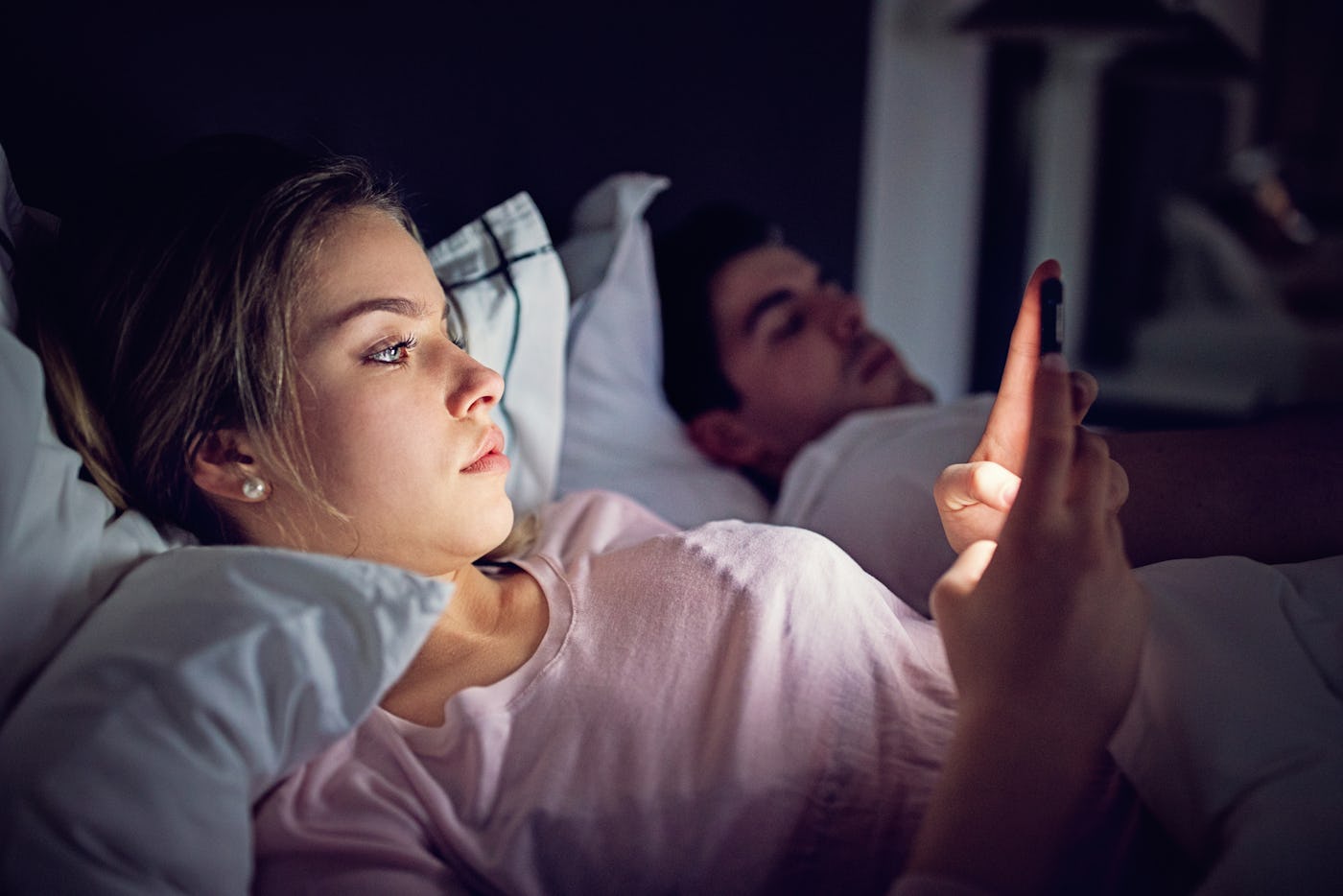 A woman scrolls on her phone in bed as her husband sleeps beside her.