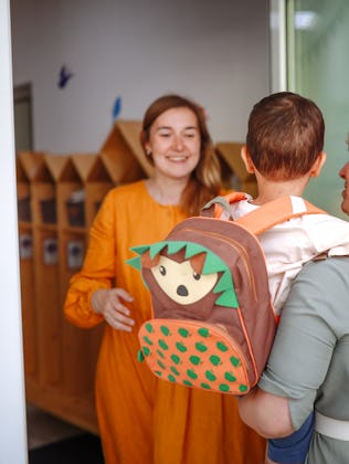 A woman carries a child with a colorful backpack into a building, greeted by another woman in an ora...