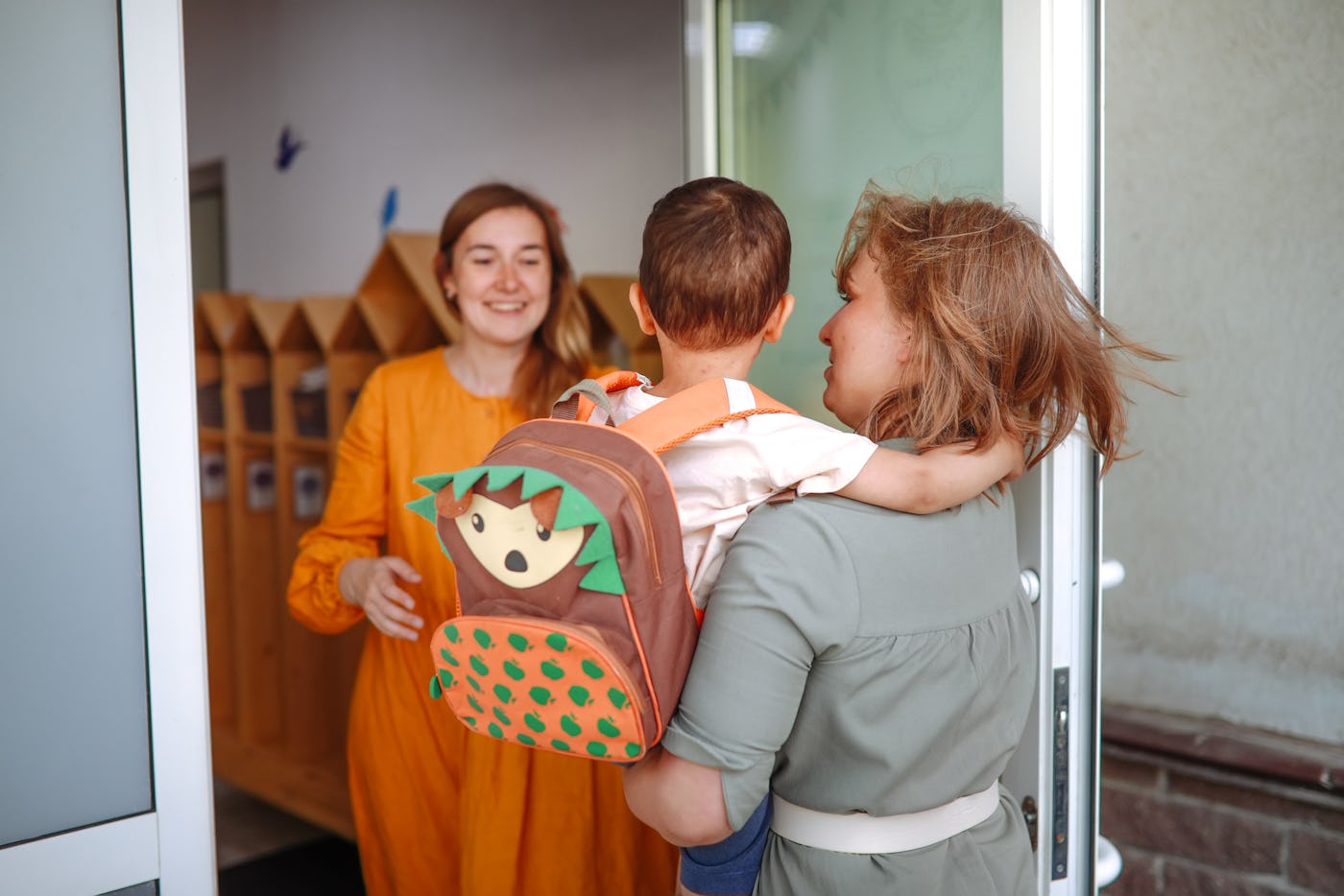 A woman carries a child with a colorful backpack into a building, greeted by another woman in an orange dress. Wooden lockers are visible inside.