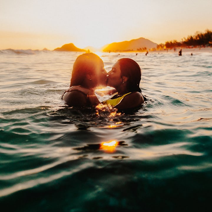 Two people embrace in the ocean at sunset, with warm golden light reflecting on the water. A distant...