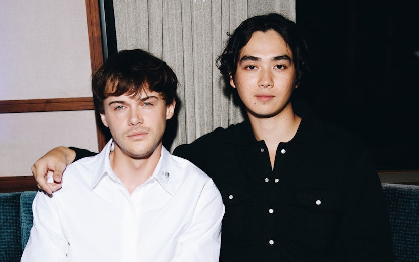 Two people sitting on a sofa indoors. One wears a white shirt and rests his arm on the other's shoul...
