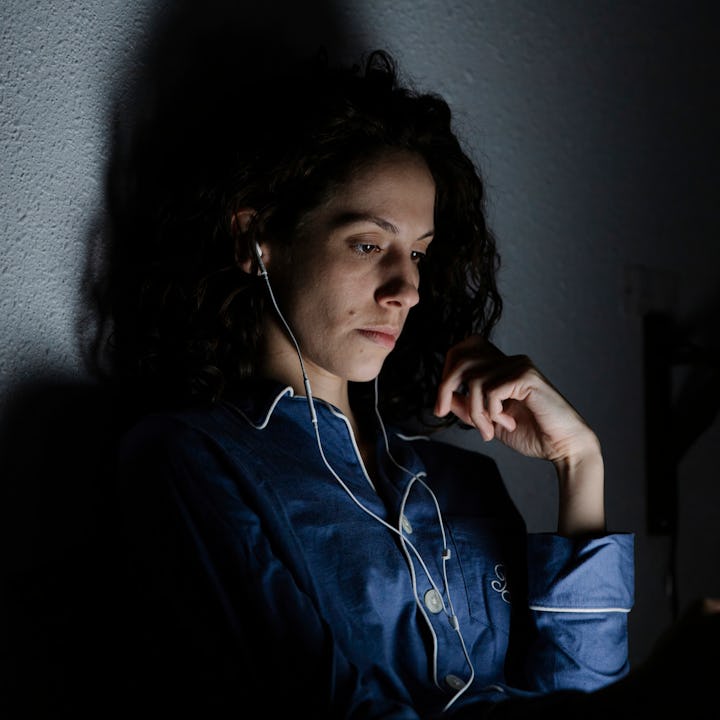 Person with curly hair wearing a blue shirt and earphones, sits in dim light, looking at a tablet sc...