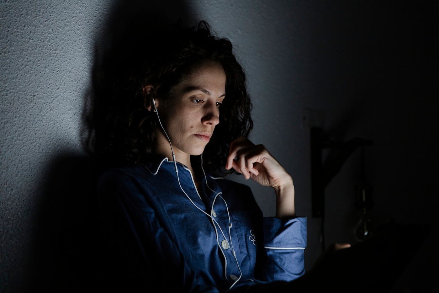 Person with curly hair wearing a blue shirt and earphones, sits in dim light, looking at a tablet screen, casting shadows on the wall behind.