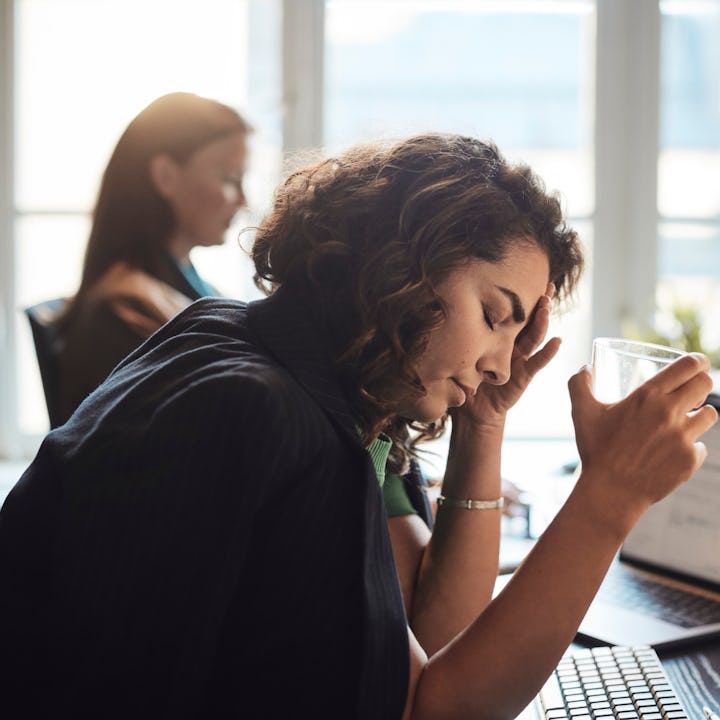 A woman sits at a desk, eyes closed, holding a glass and touching her head, looking fatigued. Anothe...