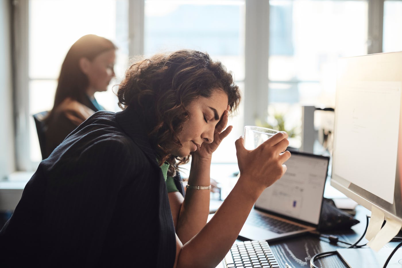 A woman sits at a desk, eyes closed, holding a glass and touching her head, looking fatigued. Another person works at a laptop in the background.
