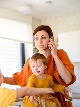 A woman in an orange shirt multitasks in a kitchen, holding a toddler and talking on the phone, whil...