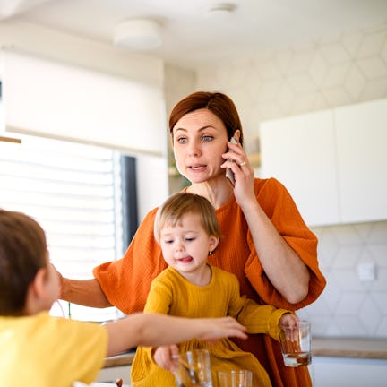 A woman in an orange shirt multitasks in a kitchen, holding a toddler and talking on the phone, whil...