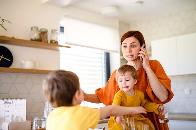 A woman in an orange shirt multitasks in a kitchen, holding a toddler and talking on the phone, whil...