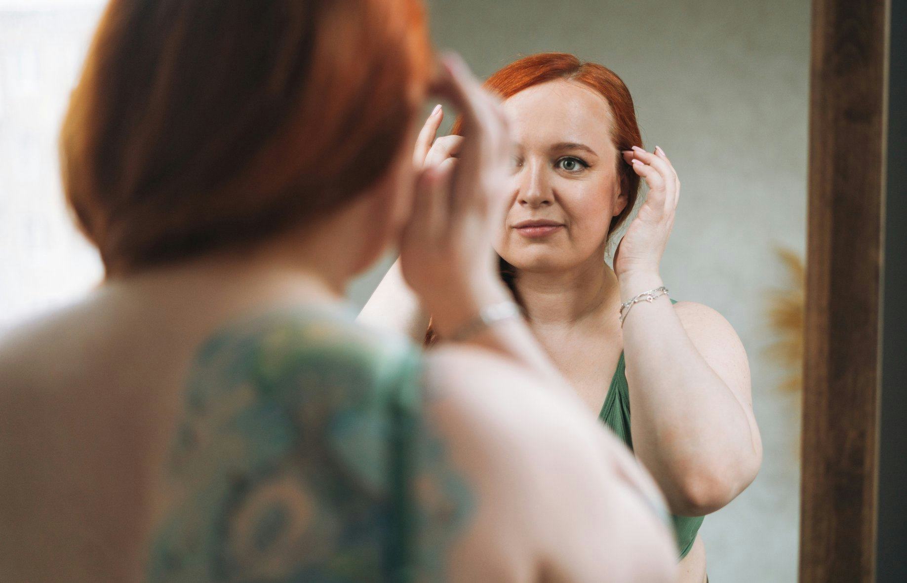 A person with red hair is adjusting their hairstyle while looking into a mirror in a softly lit room...