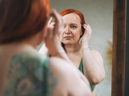 A person with red hair is adjusting their hairstyle while looking into a mirror in a softly lit room...