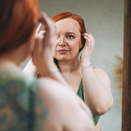 A person with red hair is adjusting their hairstyle while looking into a mirror in a softly lit room...