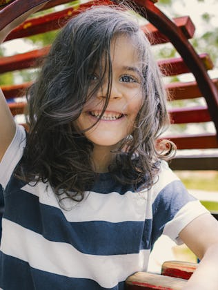 Child with long hair and a striped shirt smiling while playing on a colorful wooden playground struc...