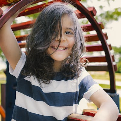 Child with long hair and a striped shirt smiling while playing on a colorful wooden playground structure. Trees and grass are visible in the background.