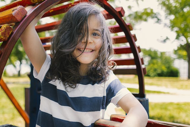 Child with long hair and a striped shirt smiling while playing on a colorful wooden playground struc...
