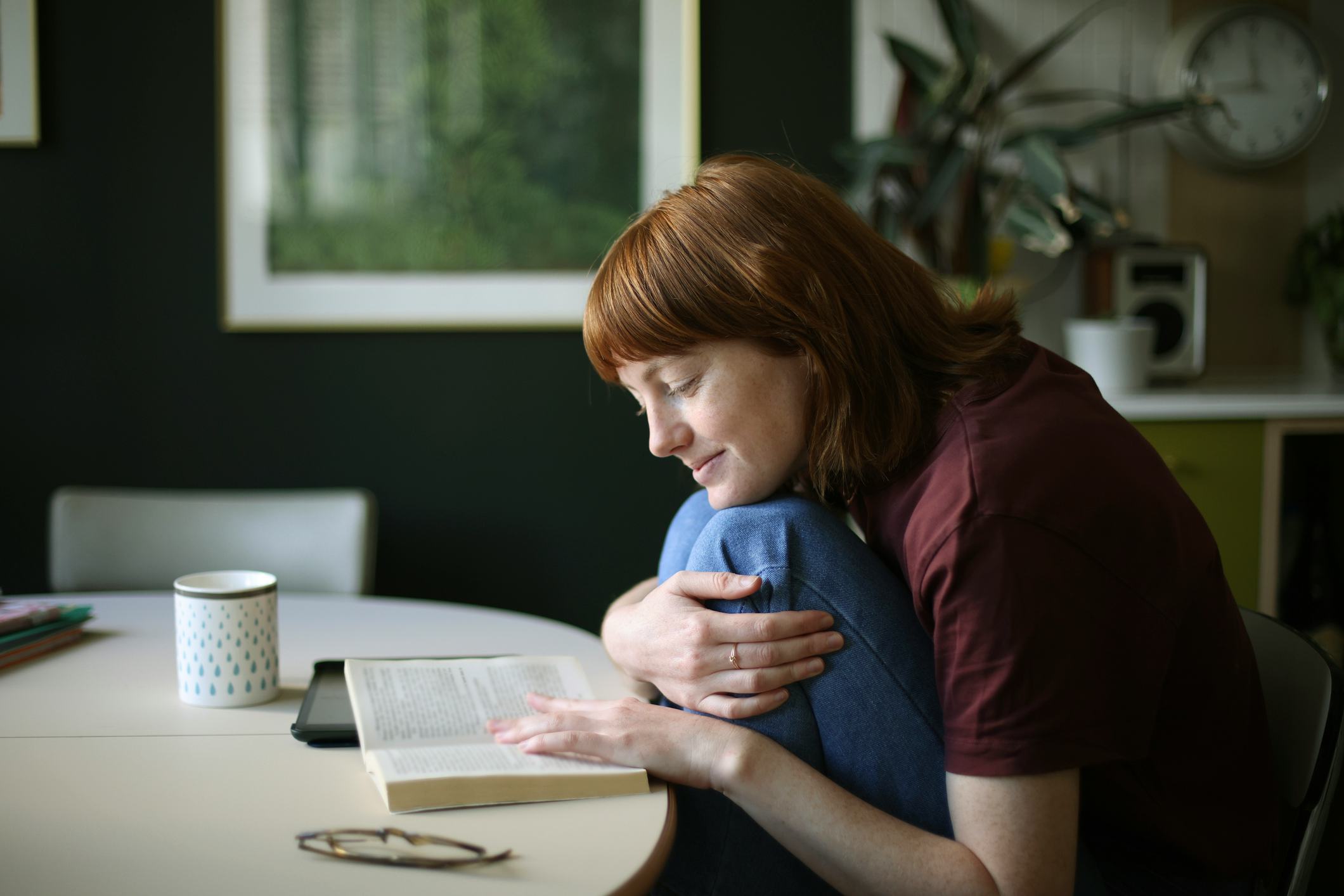 A person with red hair sits at a round table, cradling a book and smiling softly. A mug and plant ar...