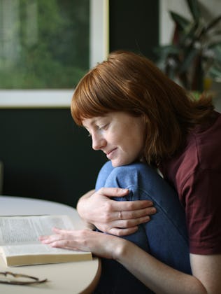 A person with red hair sits at a round table, cradling a book and smiling softly. A mug and plant ar...
