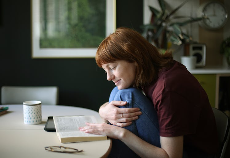 A person with red hair sits at a round table, cradling a book and smiling softly. A mug and plant ar...
