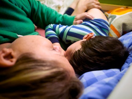A person and a child lie close together on a bed, both facing the same direction. The child is weari...