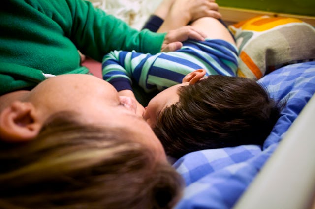 A person and a child lie close together on a bed, both facing the same direction. The child is weari...