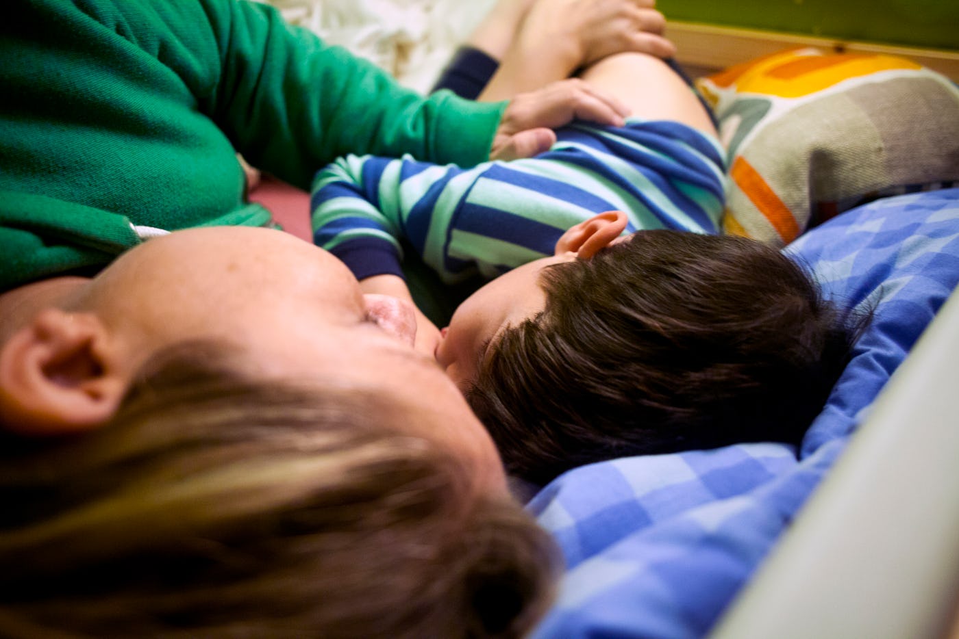 A person and a child lie close together on a bed, both facing the same direction. The child is wearing striped pajamas, and the bedding is checkered.