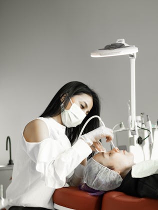 A cosmetologist wearing a mask performs a facial treatment on a client lying on a red chair, surroun...