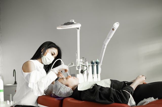 A cosmetologist wearing a mask performs a facial treatment on a client lying on a red chair, surroun...