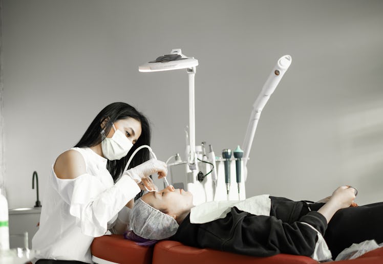 A cosmetologist wearing a mask performs a facial treatment on a client lying on a red chair, surroun...