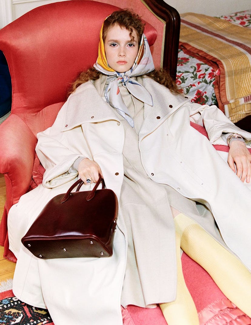 A model wearing a silk scarf tied around their head and a coat while holding a leather bag