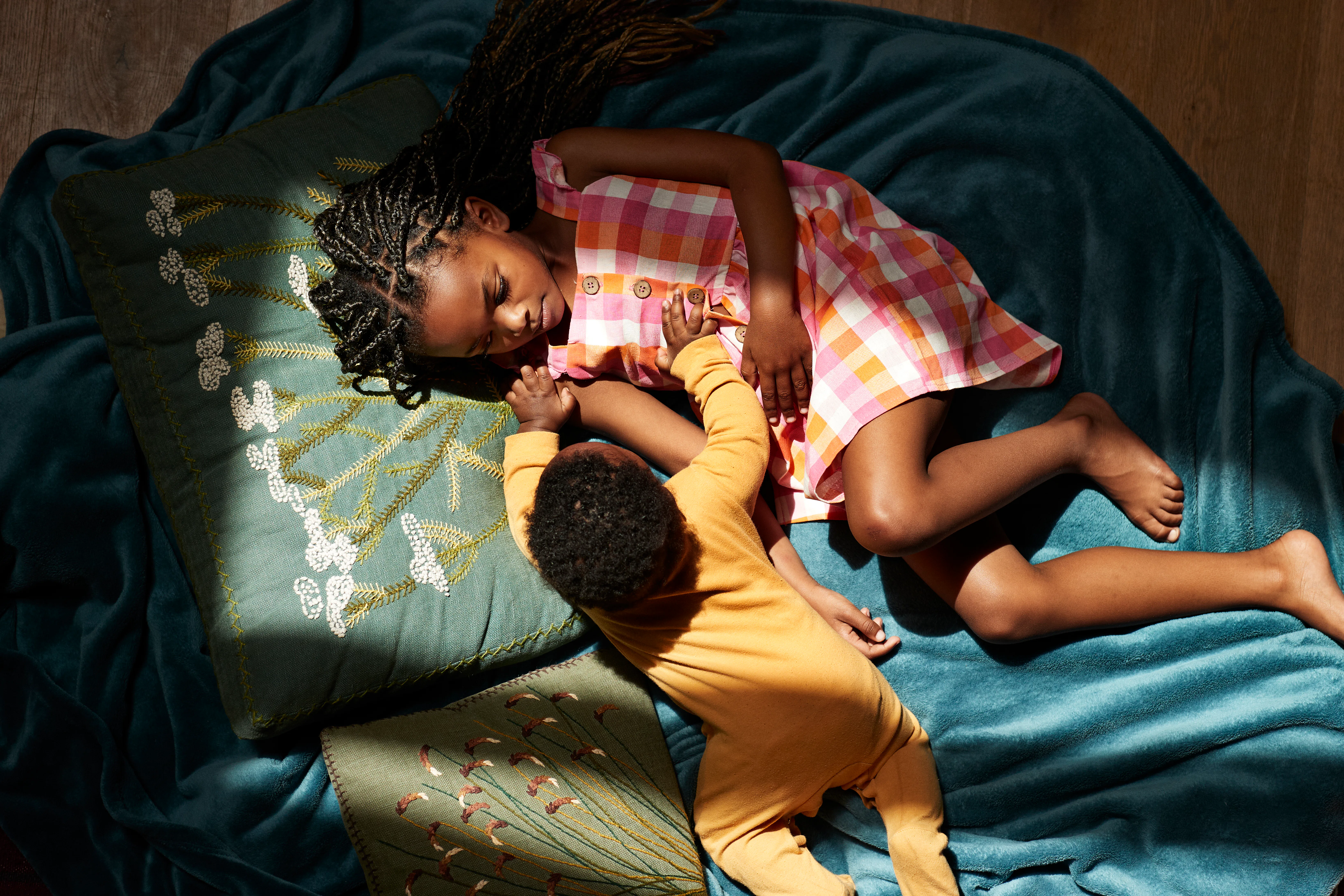 Overhead view of a girl in a pink plaid dress lying on teal fabric, holding hands with a baby in yel...