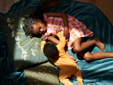 Overhead view of a girl in a pink plaid dress lying on teal fabric, holding hands with a baby in yel...