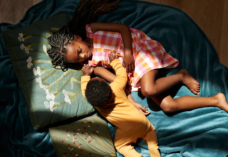 Overhead view of a girl in a pink plaid dress lying on teal fabric, holding hands with a baby in yel...