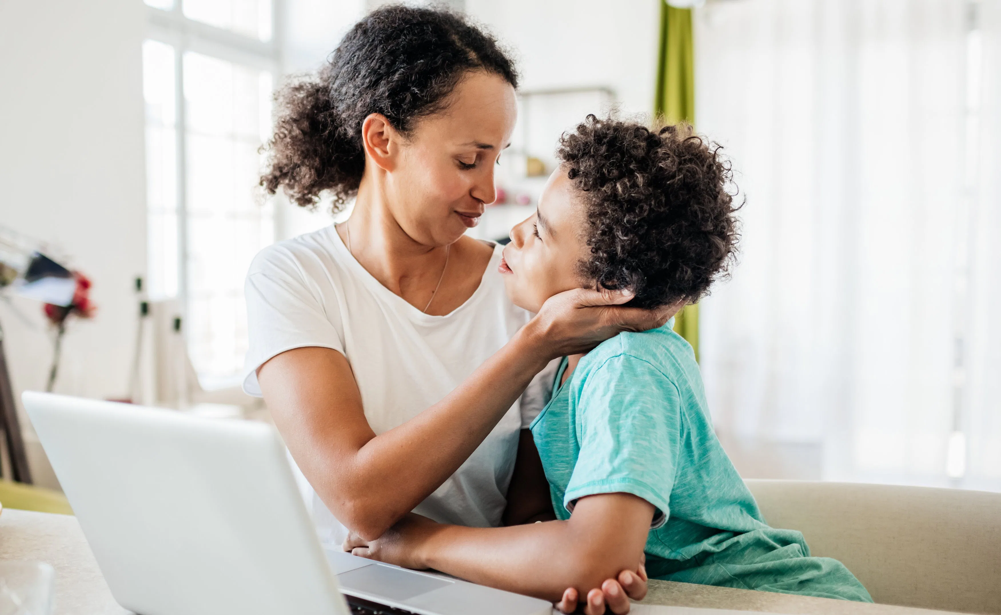 A mother gently holds her young son’s face as they share a close, affectionate moment at a table wit...