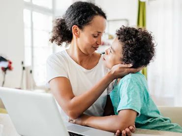 A mother gently holds her young son’s face as they share a close, affectionate moment at a table wit...
