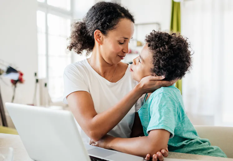A mother gently holds her young son’s face as they share a close, affectionate moment at a table wit...