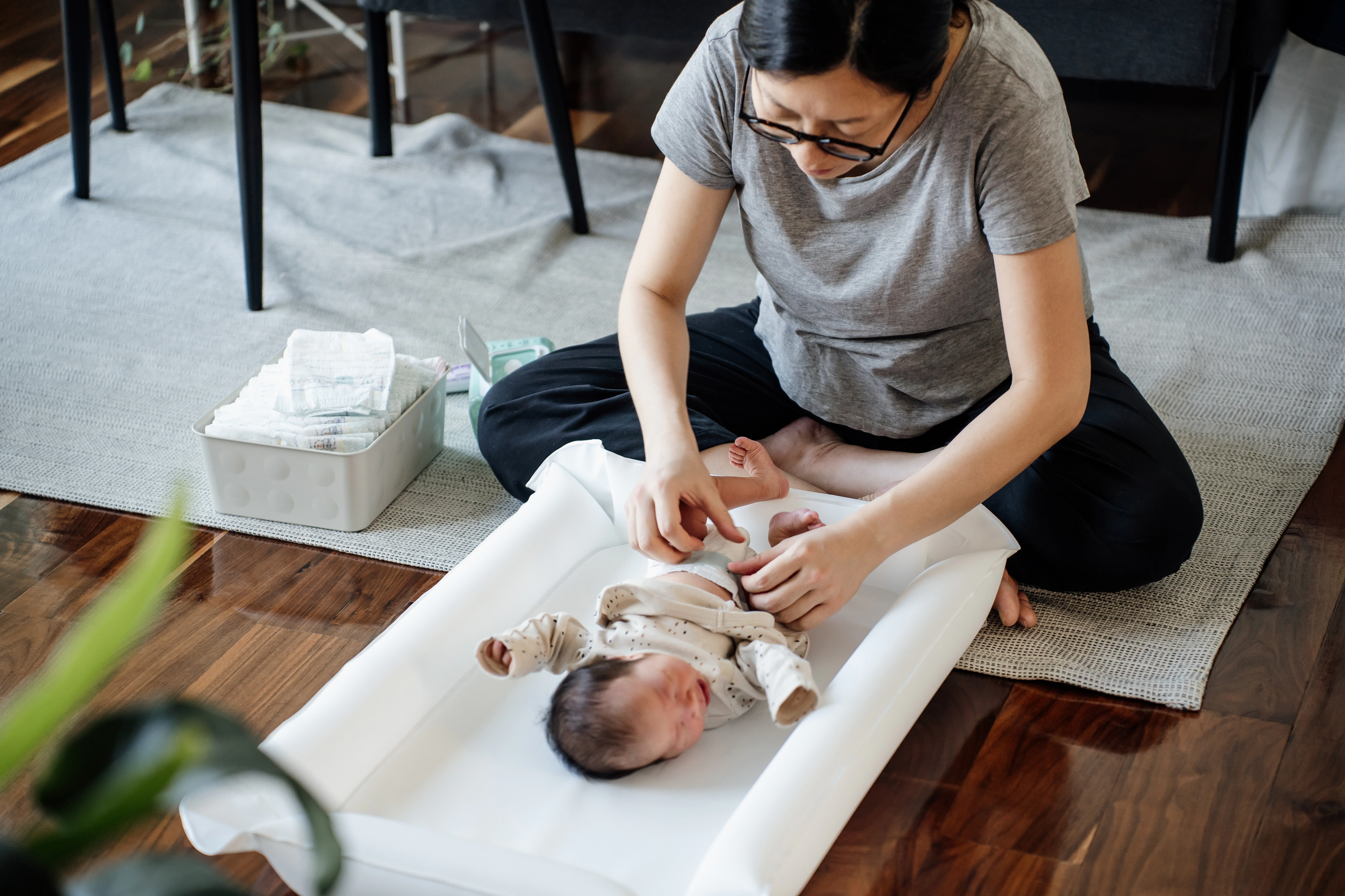 A seated parent gently dressing a newborn on a white changing pad on the floor, with diaper supplies...