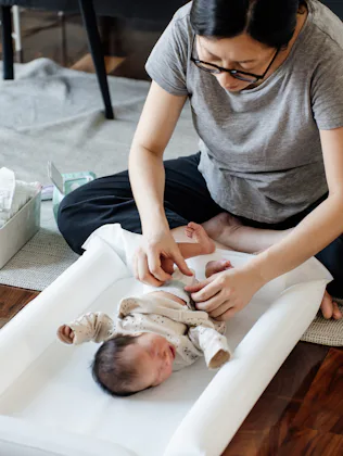 A seated parent gently dressing a newborn on a white changing pad on the floor, with diaper supplies...