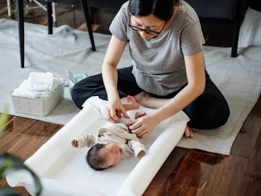 A seated parent gently dressing a newborn on a white changing pad on the floor, with diaper supplies...
